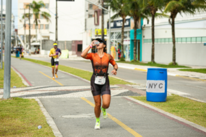 Female runner hydrating during a race, wearing athletic gear and drinking water while running on a sunny road surrounded by other racers.